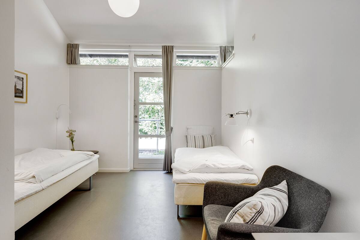 Una habitación minimalista con dos camas, un sofá gris, una mesita de noche con lámpara y una ventana con cortinas.