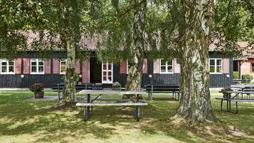 A picnic area with benches and picnic tables, surrounded by trees and a building with red shutters in the background.