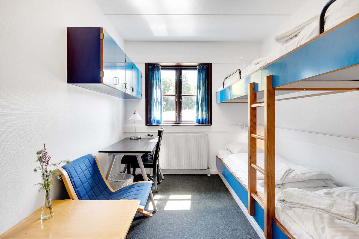Habitación tipo dormitorio con literas, escritorio, silla, radiador y ventana con cortinas.
