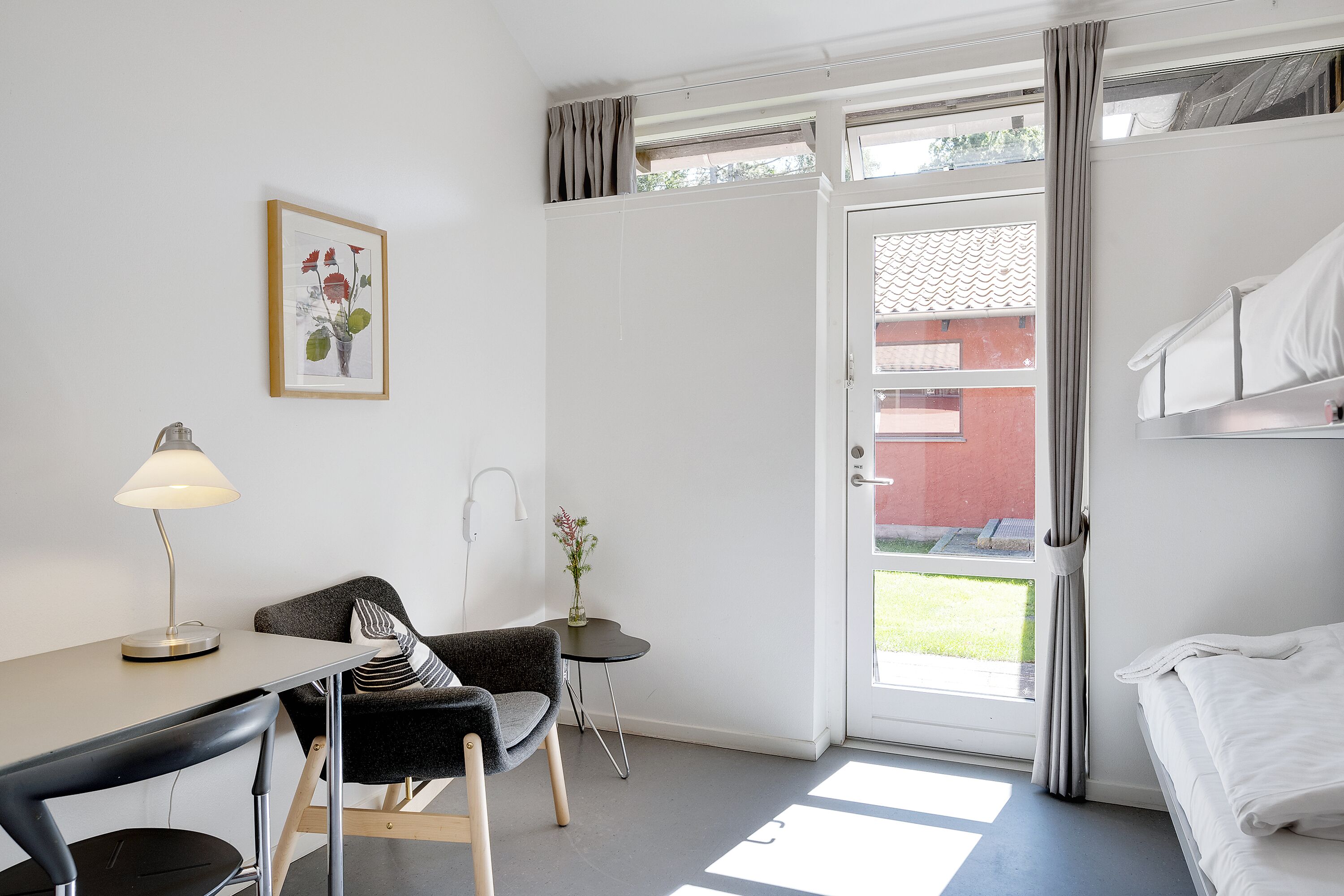 A room with a desk, chair, bed, and a small table with a vase.