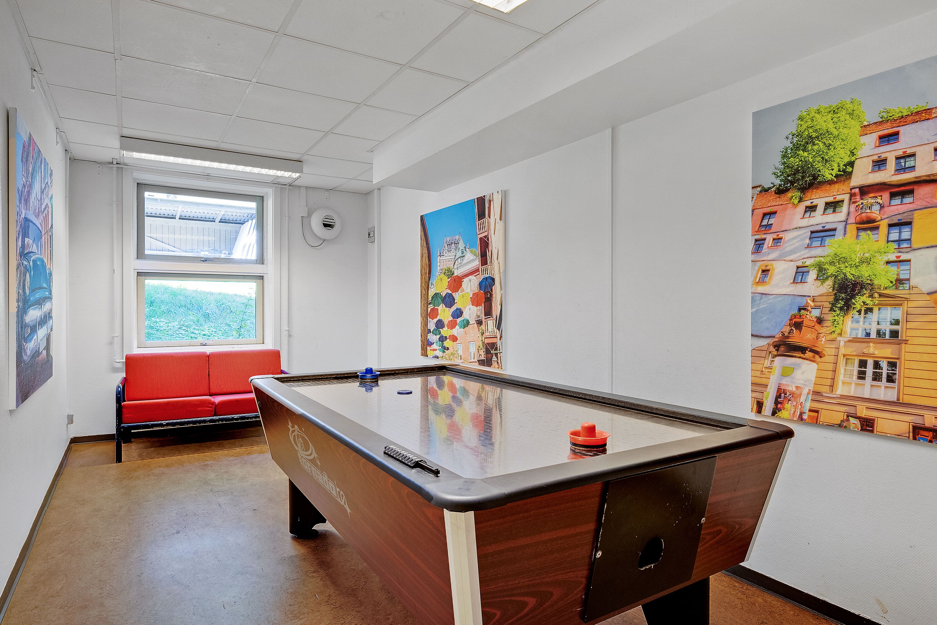 Una mesa de billar en una habitación con un sofá rojo, una ventana y coloridas obras de arte en las paredes.