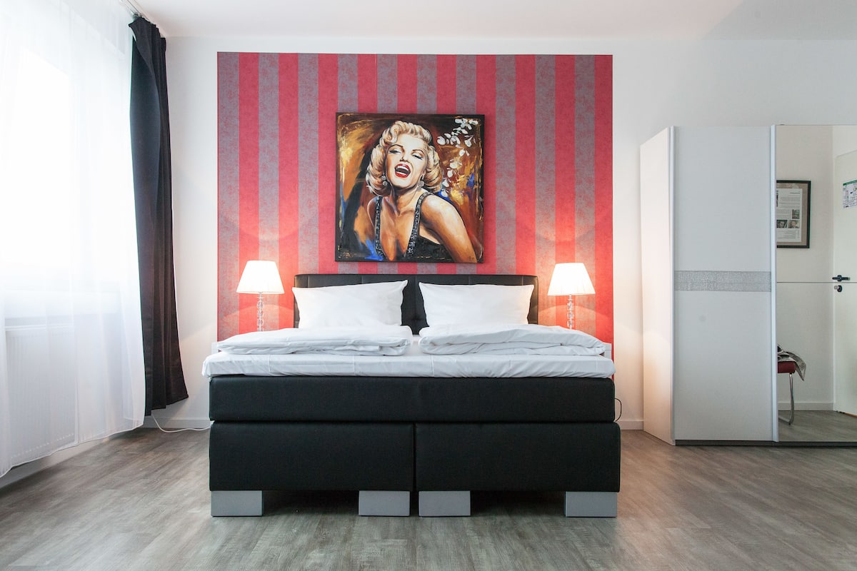A modern hotel room with a large bed, two bedside lamps, a red-striped wall, and a painting above the bed.