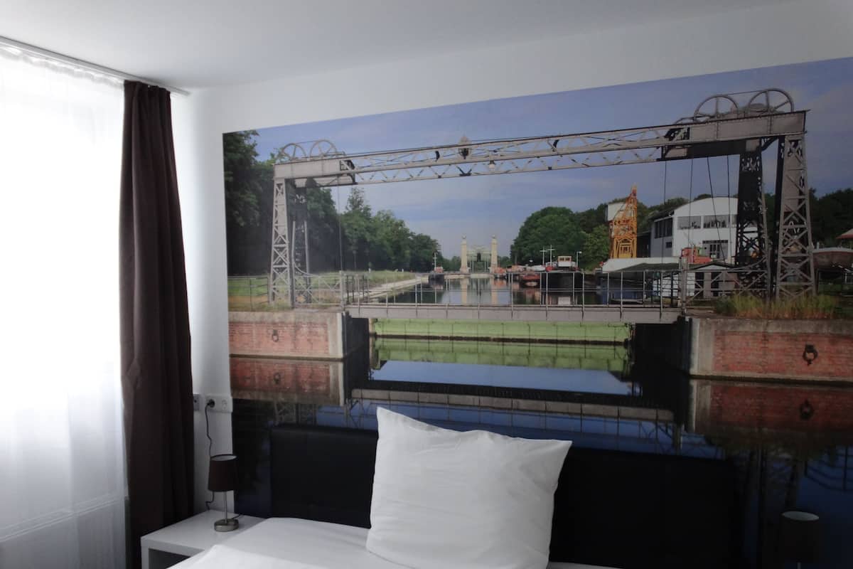 A hotel room with a bed, a nightstand, a lamp, and a large mural of a bridge and water.