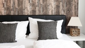 Un lit avec des oreillers blancs et gris, une tête de lit en bois et une table de chevet sur laquelle se trouvent une lampe et une pile de bûches.