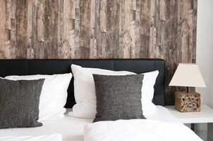 Una cama con almohadas blancas y grises, un cabecero de madera y una mesita de noche con una lámpara y una pila de leños.