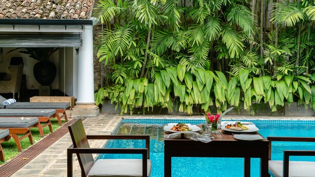 Un espace aménagé au bord de la piscine, avec des chaises longues, une table à manger dressée et un décor luxuriant de verdure en arrière-plan.