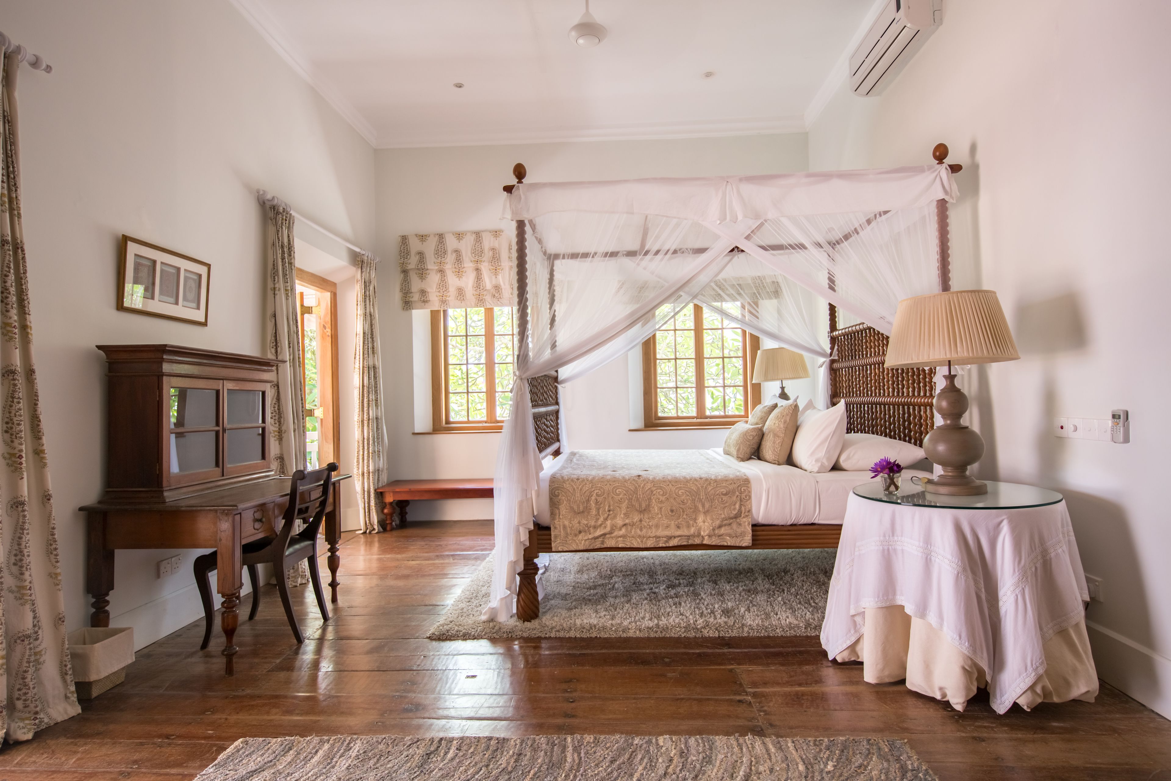 A bedroom with a four-poster bed, a wooden desk, and a bedside table with a lamp.