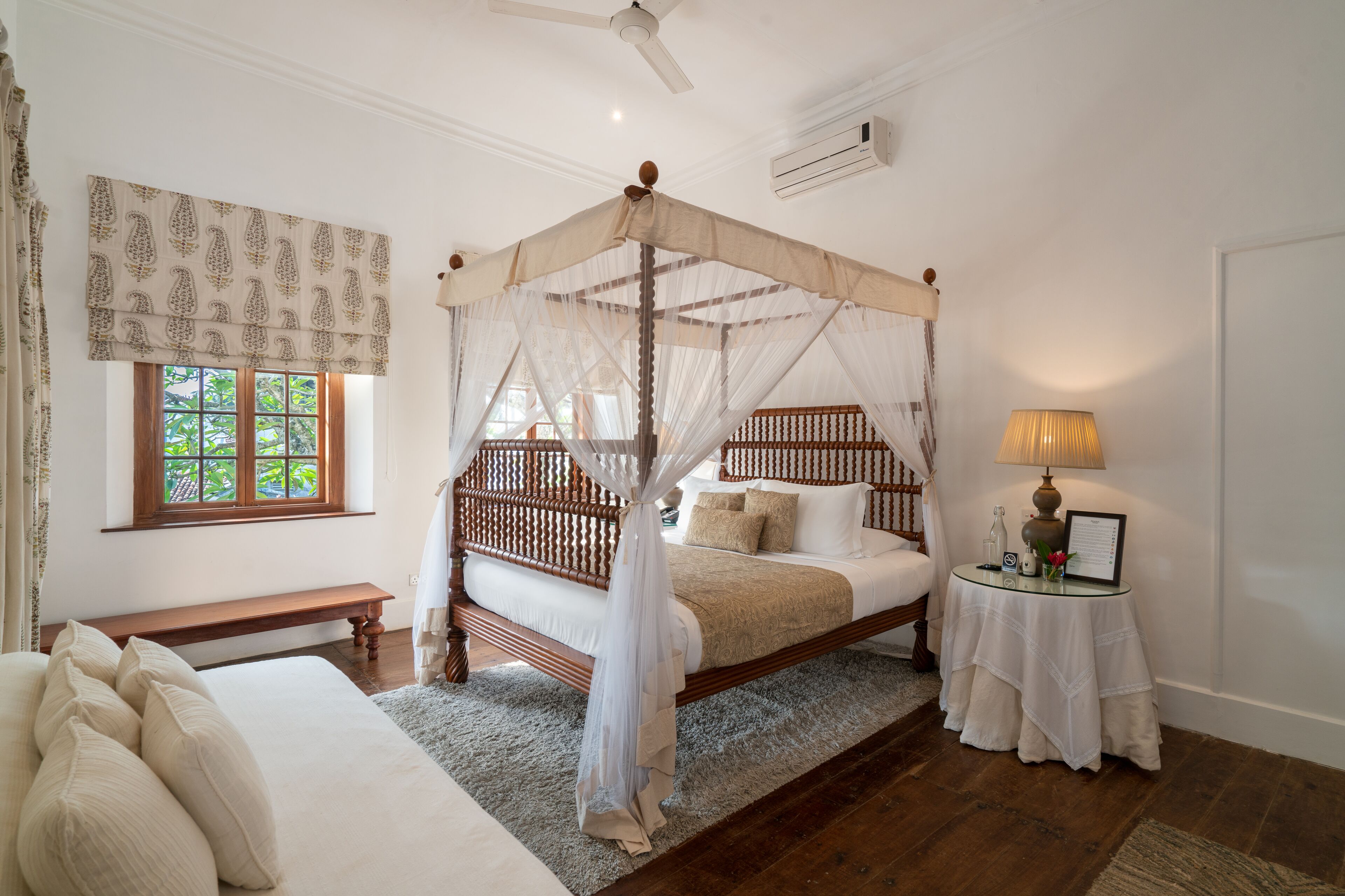 A bedroom with a four-poster canopy bed, a bedside table with a lamp, a bench, and a window with patterned curtains.