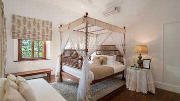 A bedroom with a four-poster canopy bed, a bedside table with a lamp, a bench, and a window with patterned curtains.