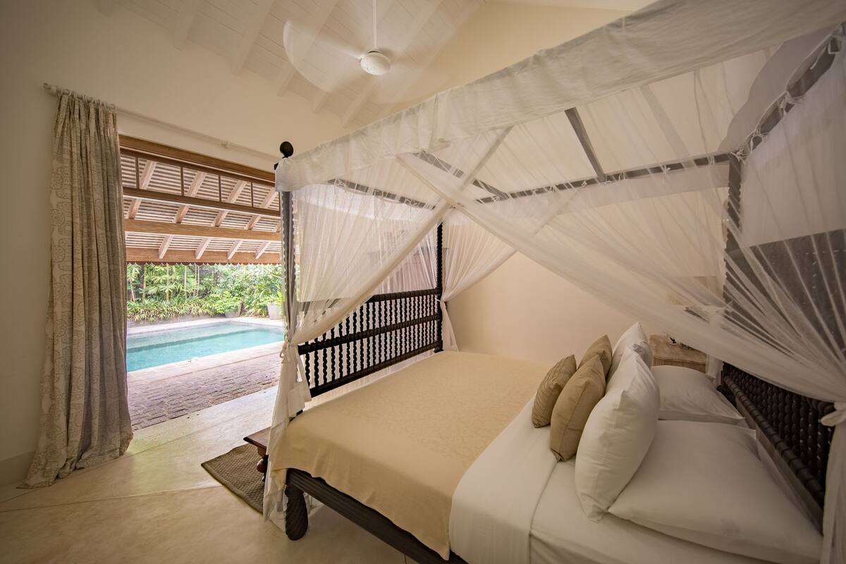 Un dormitorio con cama bajo dosel, vista a una piscina y un ventilador de techo de madera.