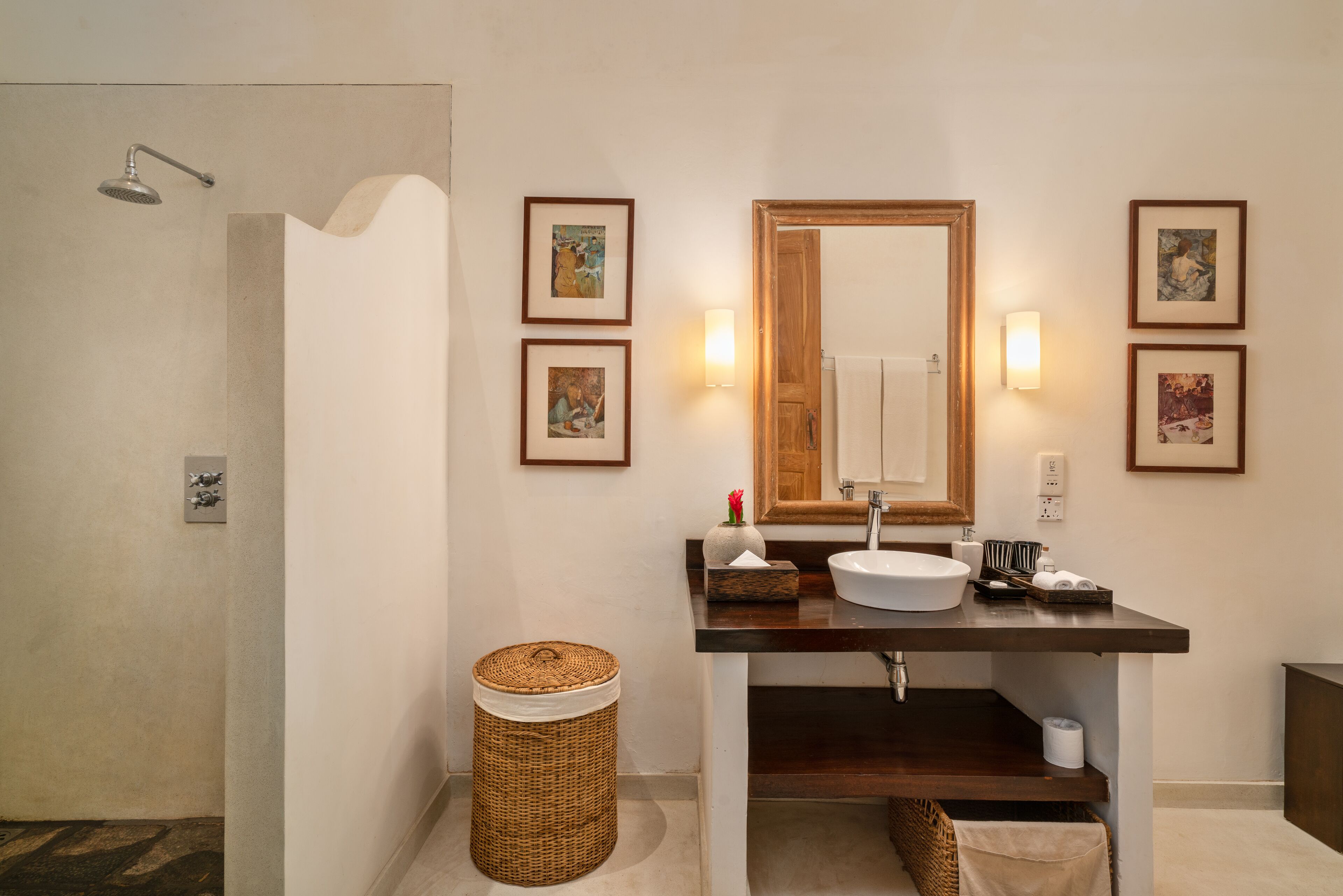 A bathroom with a shower, sink, and mirror. There are framed pictures on the wall and a trash can.