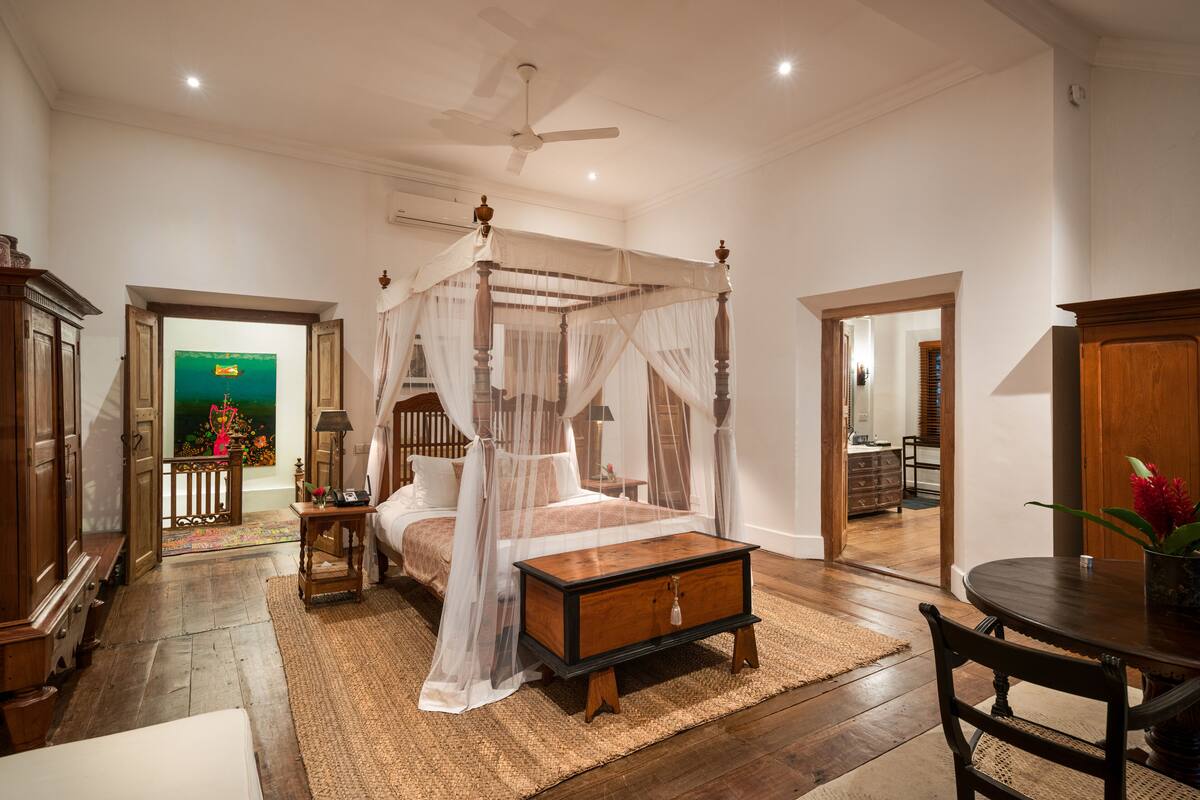 Un dormitorio espacioso con una cama con dosel, una mesita de noche de madera, una mesilla auxiliar y un ventanal grande.