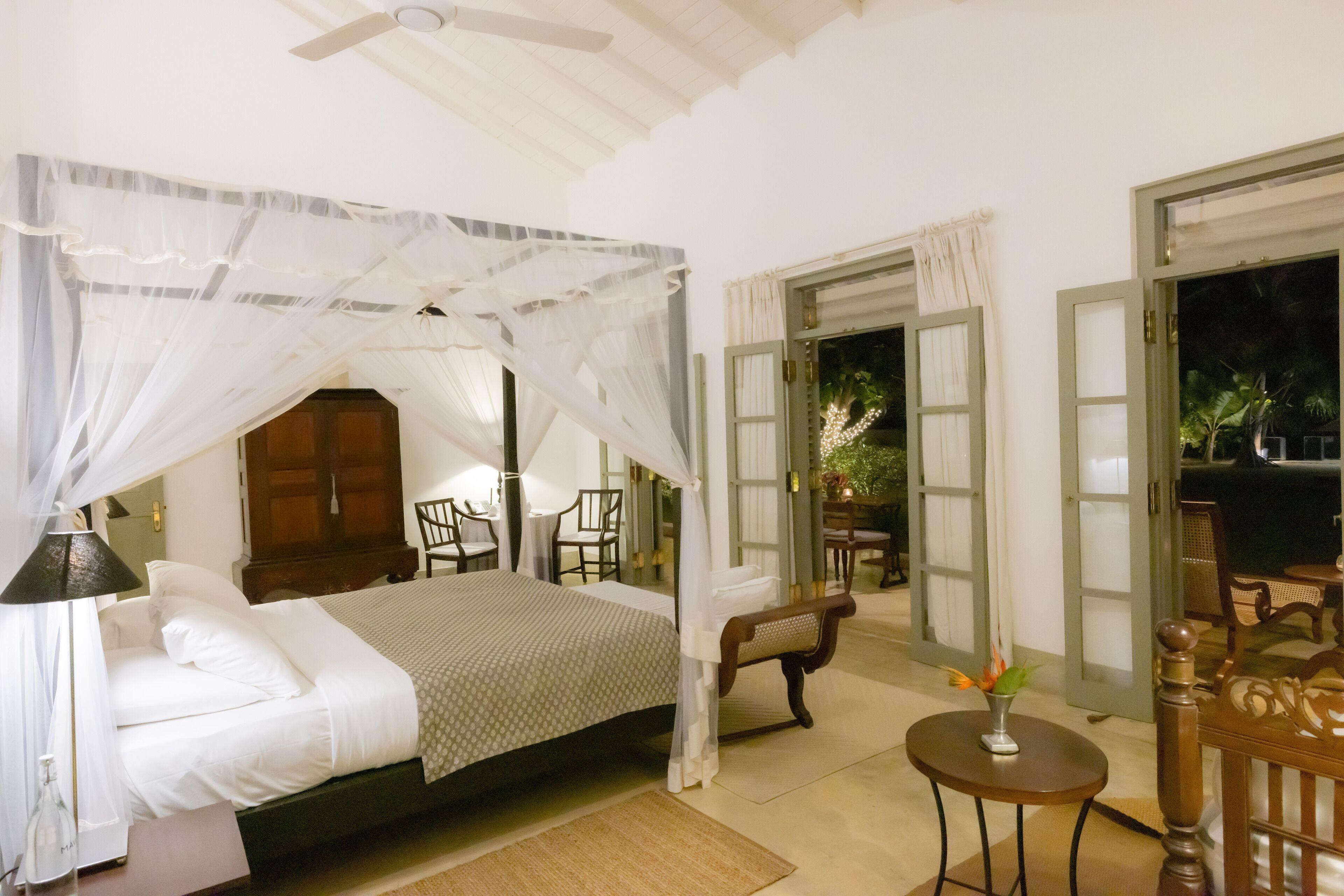 A bedroom with a canopy bed, a bedside table, a chair, and a small table with a vase. There is a door leading to an outdoor area with chairs and a table.