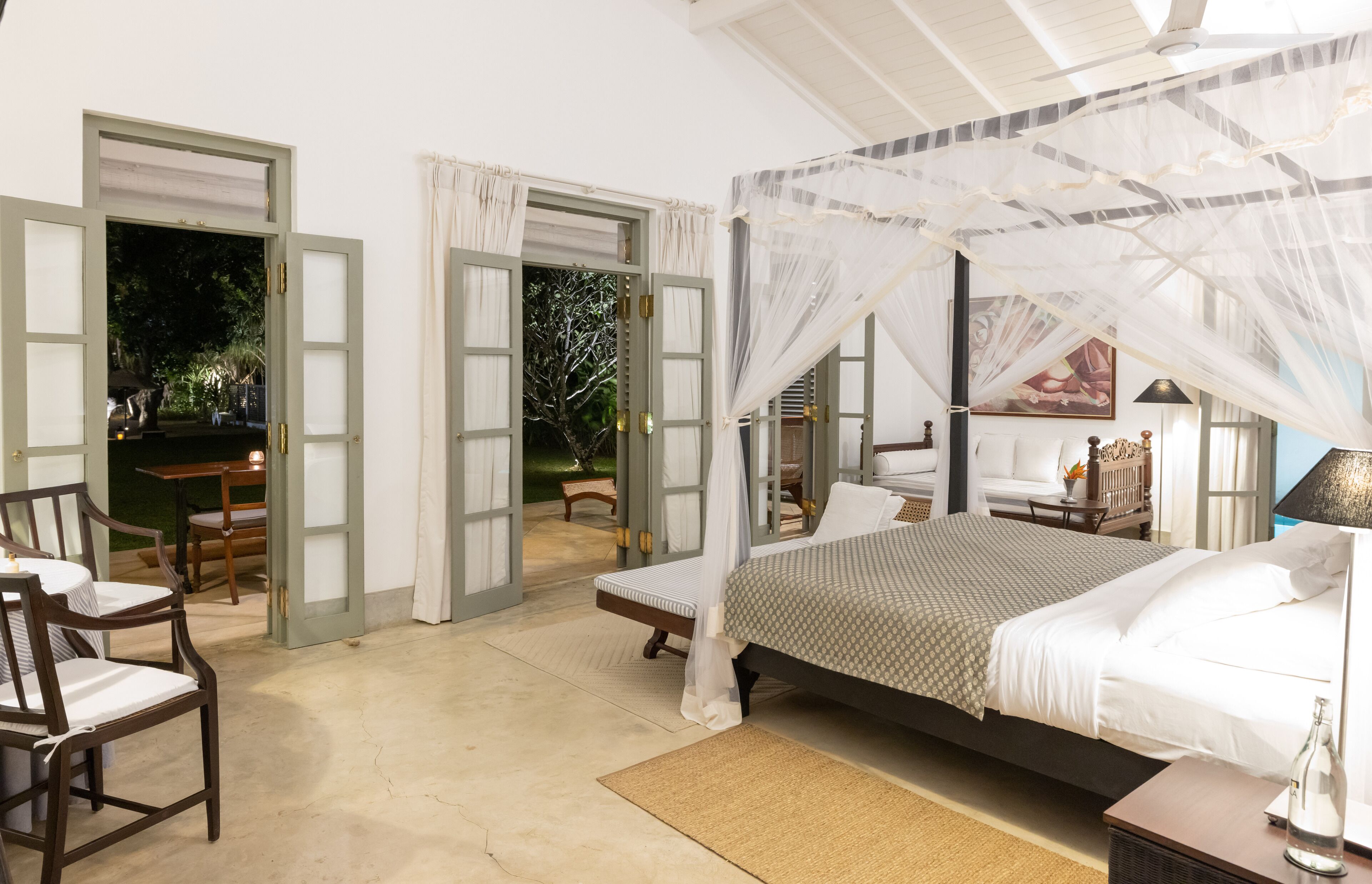 A spacious bedroom with a large bed, a sitting area with chairs, and a view of the garden through open doors.