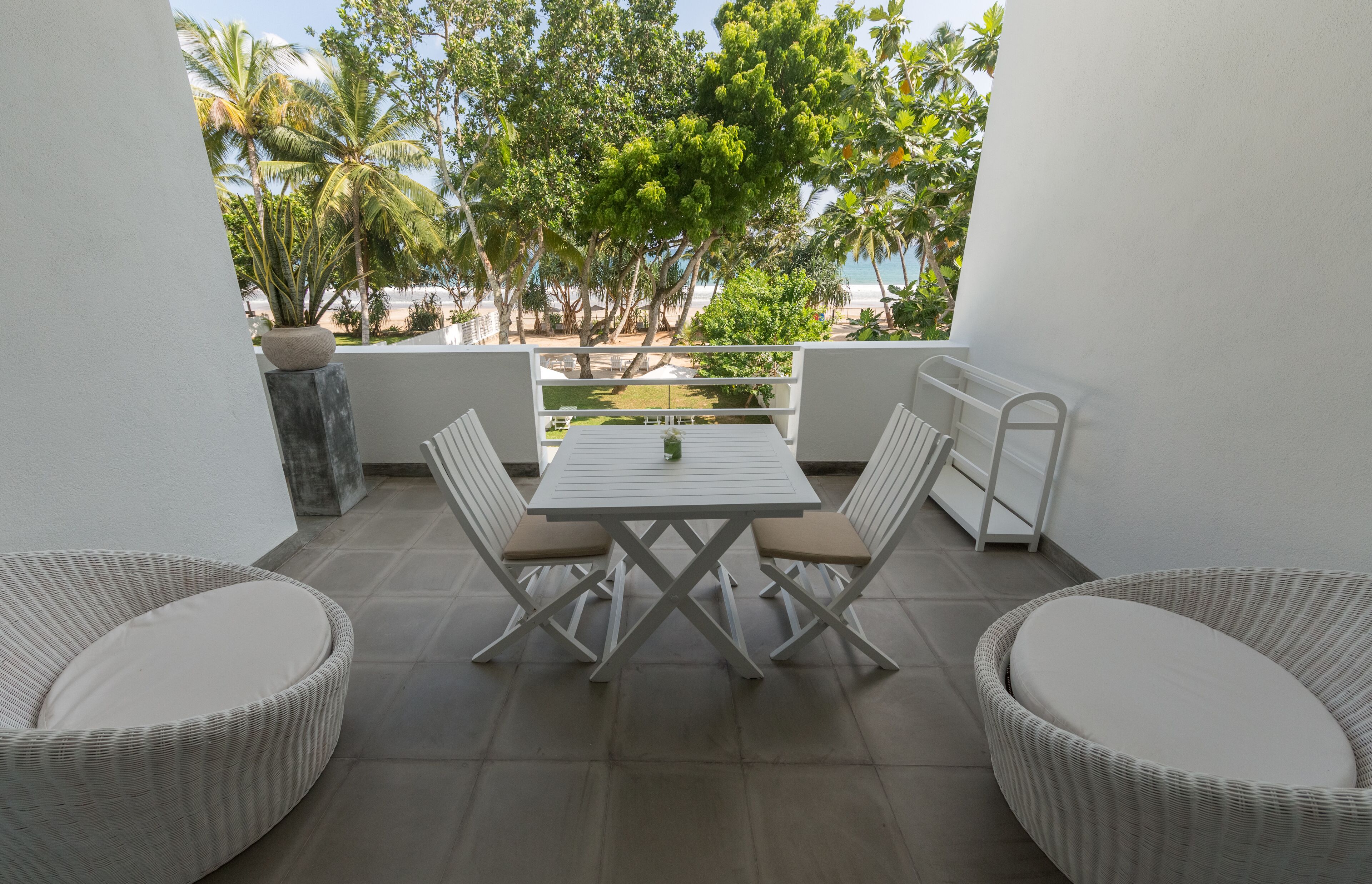 A balcony with a white table and chairs, a potted plant, and a view of a beach and palm trees.