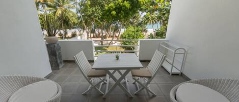 A balcony with a white table and chairs, a potted plant, and a view of a beach and palm trees.