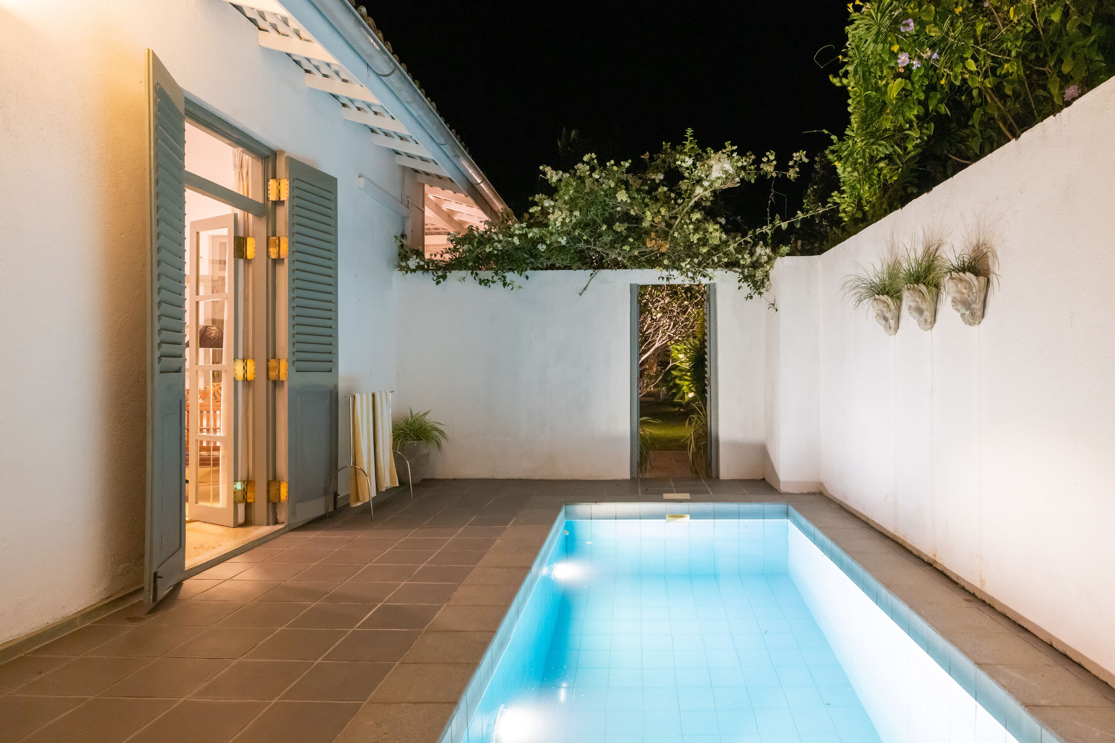 A swimming pool with a tiled deck, a white wall with hanging planters, and a door leading to an interior space.