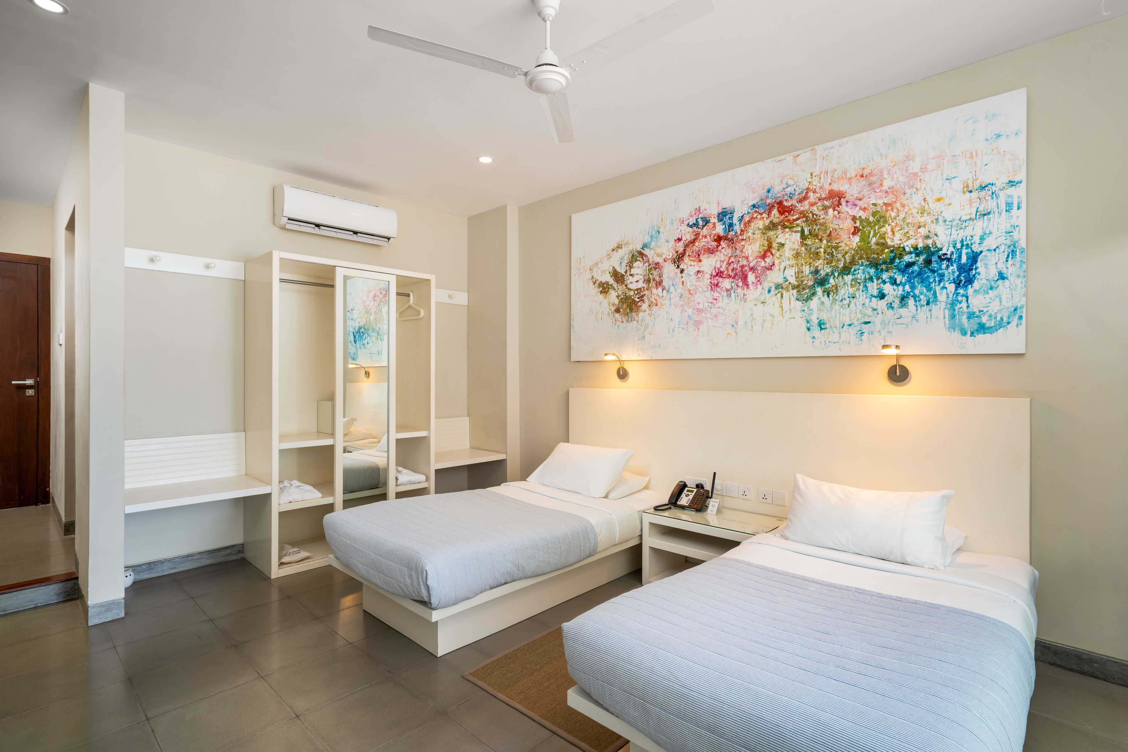 A hotel room with two beds, a wardrobe, a ceiling fan, an air conditioning unit, and a large abstract painting on the wall.