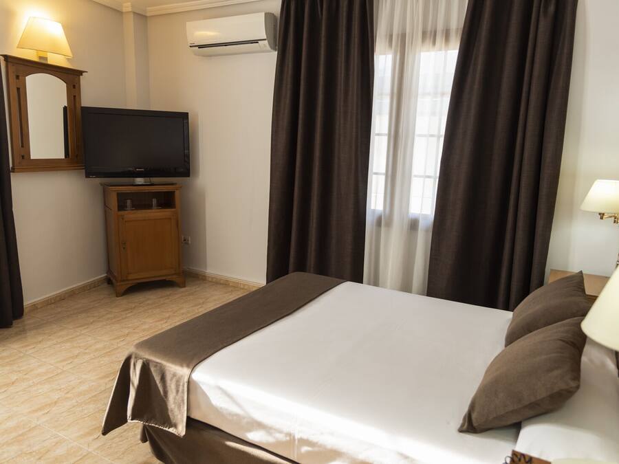 A hotel room with a bed, a television on a wooden cabinet, an air conditioning unit, and a window with curtains.