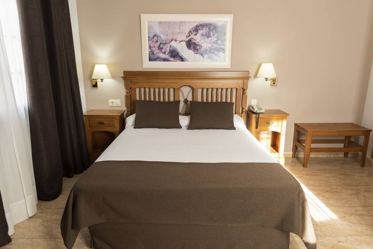 A hotel room with a large bed, two bedside tables with lamps, a wooden nightstand, and a painting on the wall.