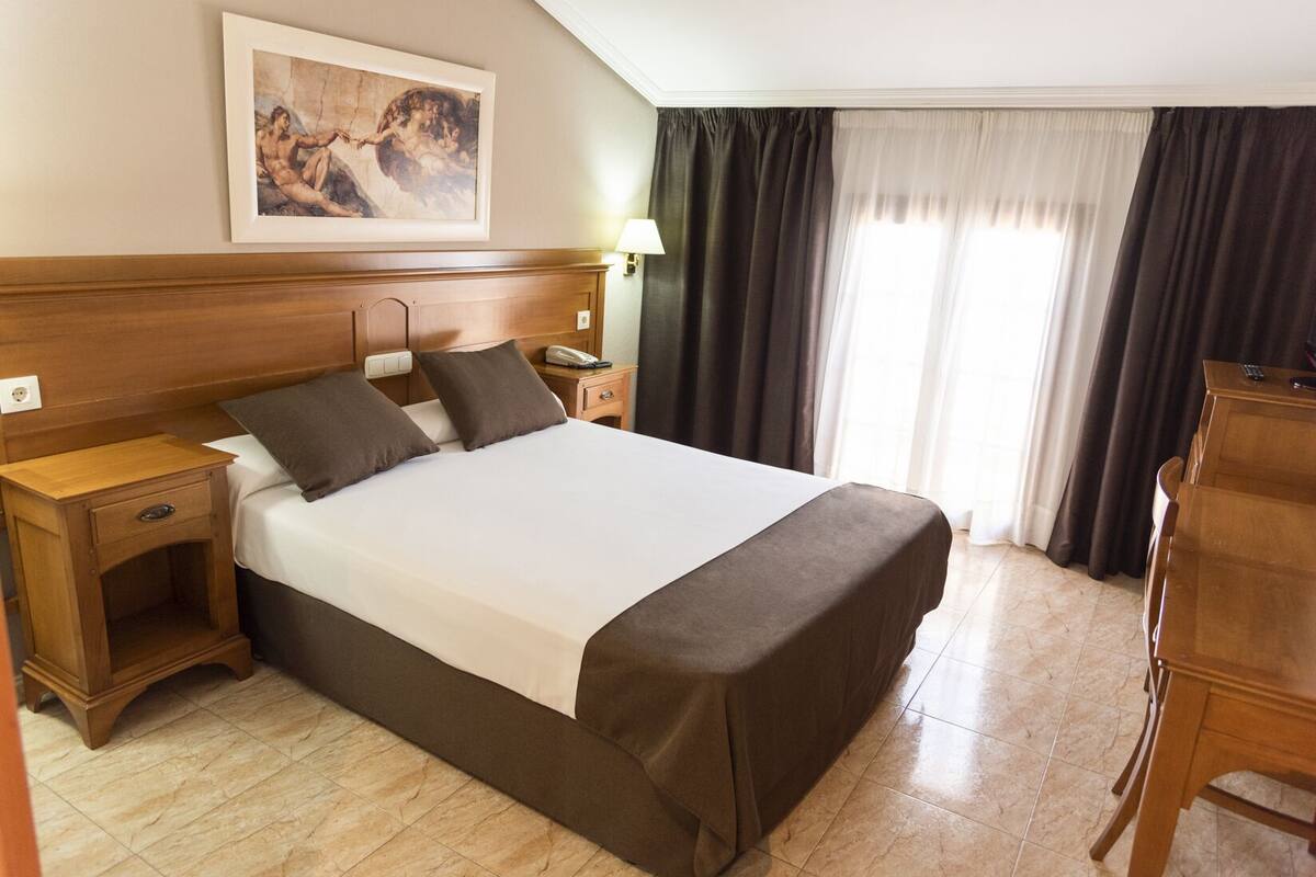 A hotel room with a large bed, wooden headboard, a nightstand with a lamp, a desk, and a chair.