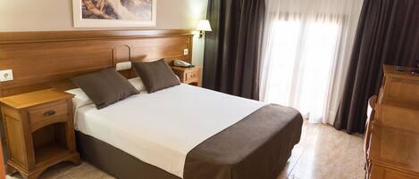 Habitación de hotel con una cama grande, cabecero de madera, mesita de noche con lámpara, escritorio y una silla.