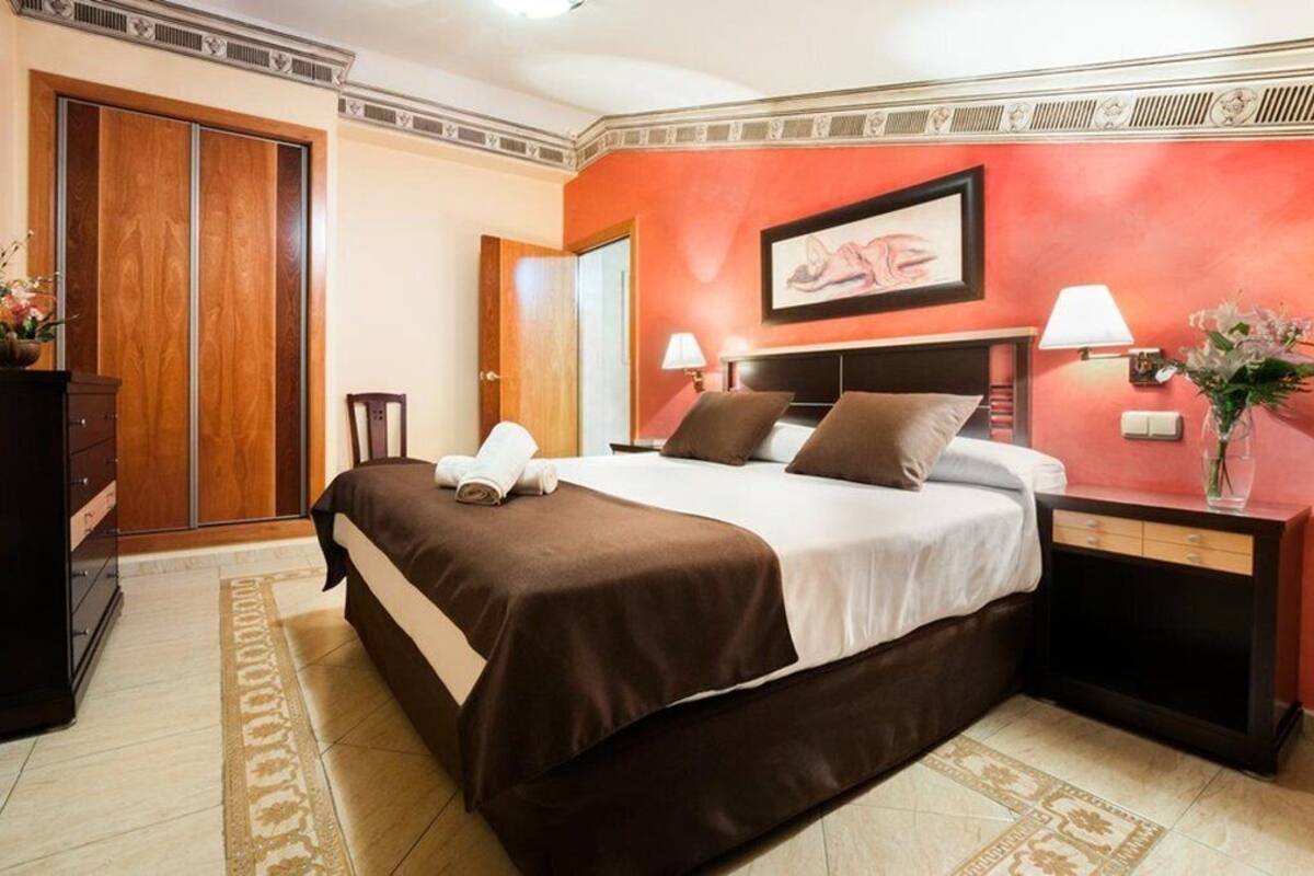 A hotel room with a large bed, a wooden wardrobe, a nightstand with a lamp, and a framed artwork on the wall.