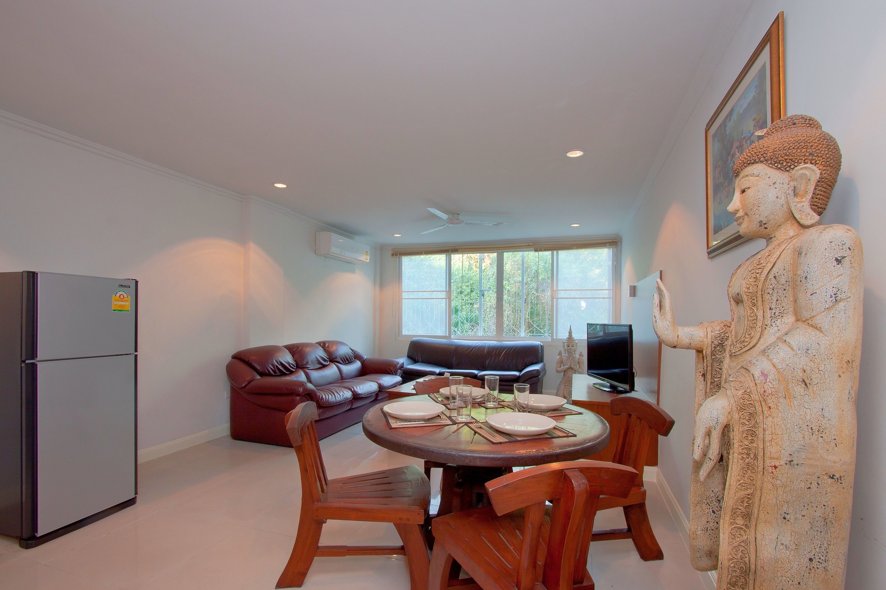A dining area with a round table, chairs, a refrigerator, a sofa, and a Buddha statue.