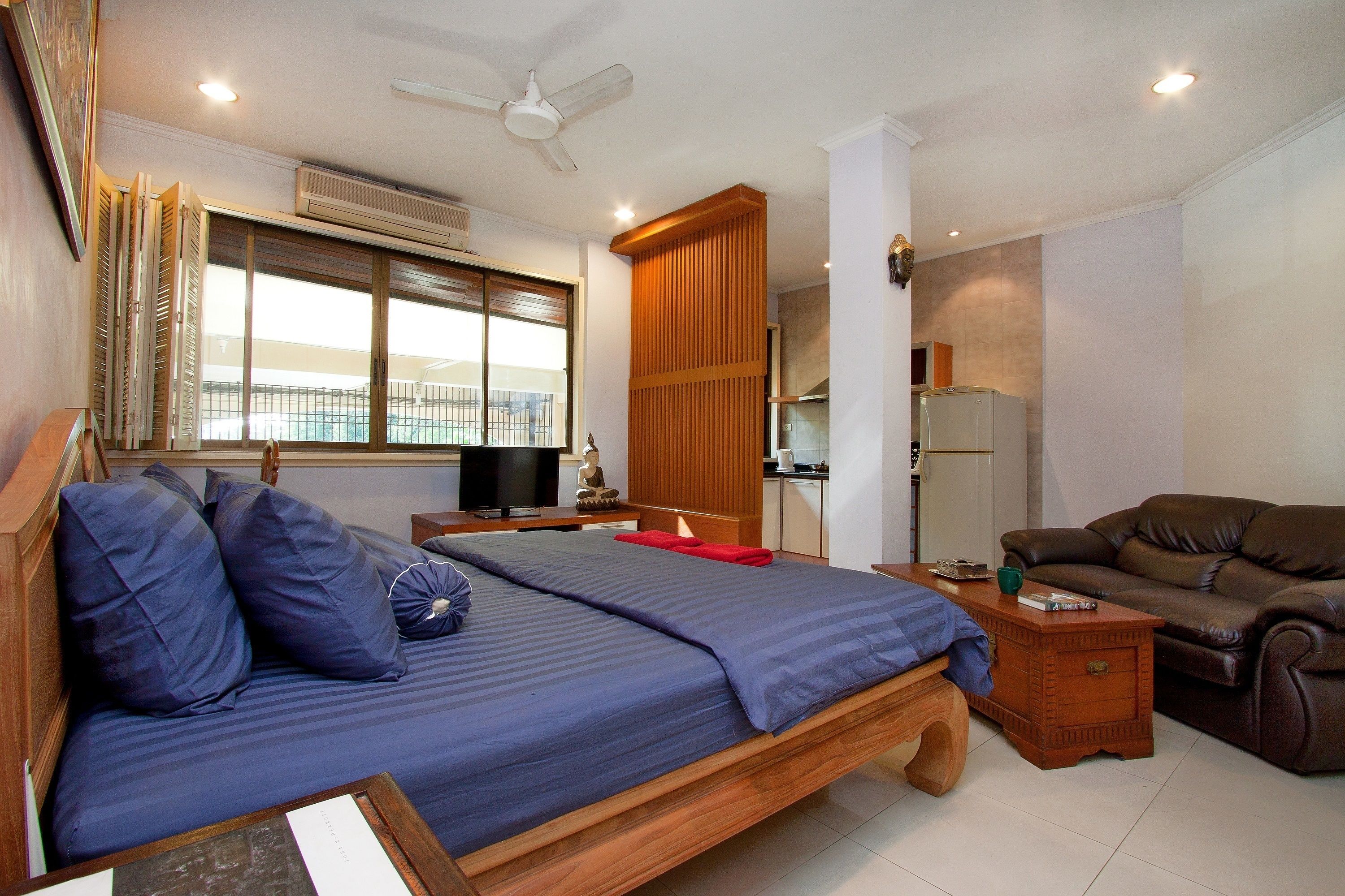 A bedroom with a bed, a wooden nightstand, a brown leather sofa, a ceiling fan, and a window with blinds.