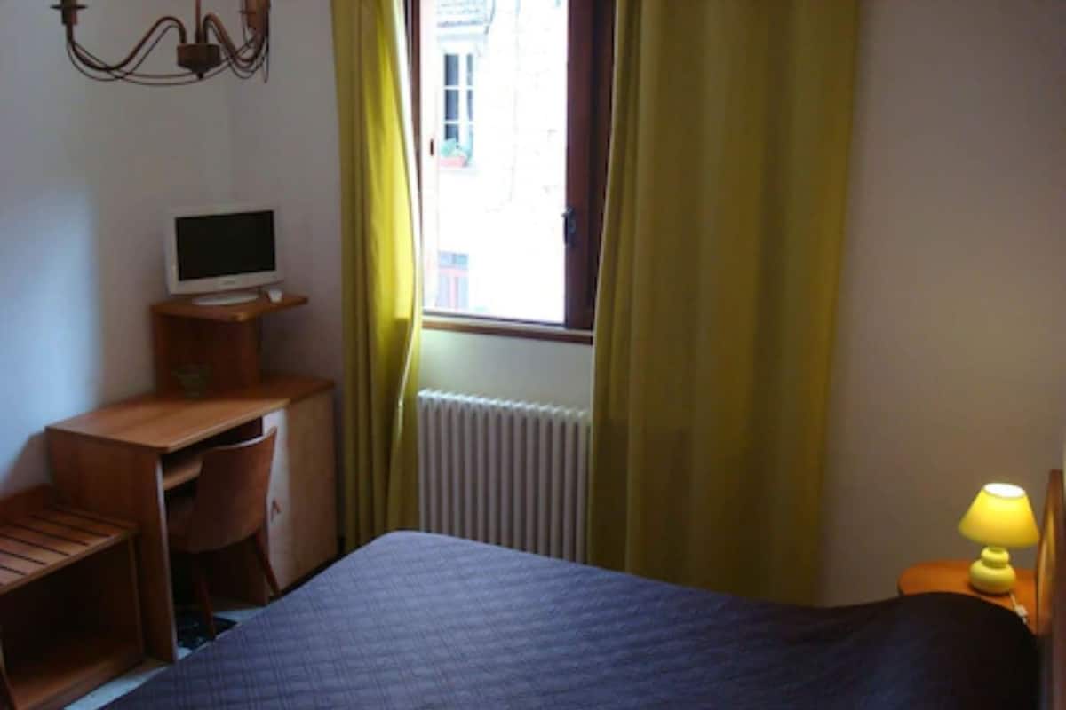 A hotel room with a bed, a desk with a computer, a lamp, and a window with curtains.