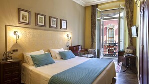 Habitación de hotel con una cama grande, una mesita de noche con teléfono, una silla y un balcón con vista a un edificio.