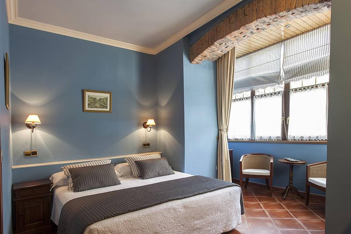 A bedroom with a large bed, two bedside lamps, a window with curtains, a small table, and a chair.
