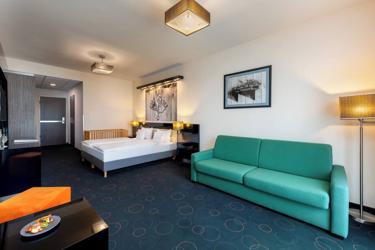 A hotel room with a bed, a green sofa, a desk with a fruit plate, a lamp, and a painting on the wall.