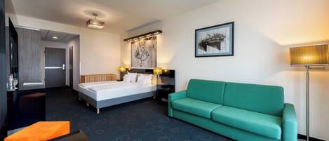A hotel room with a bed, a green sofa, a desk with a fruit plate, a lamp, and a painting on the wall.