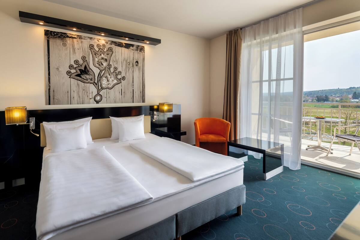 A hotel room with a large bed, two bedside tables with lamps, a chair, and a view of the countryside through a large window.