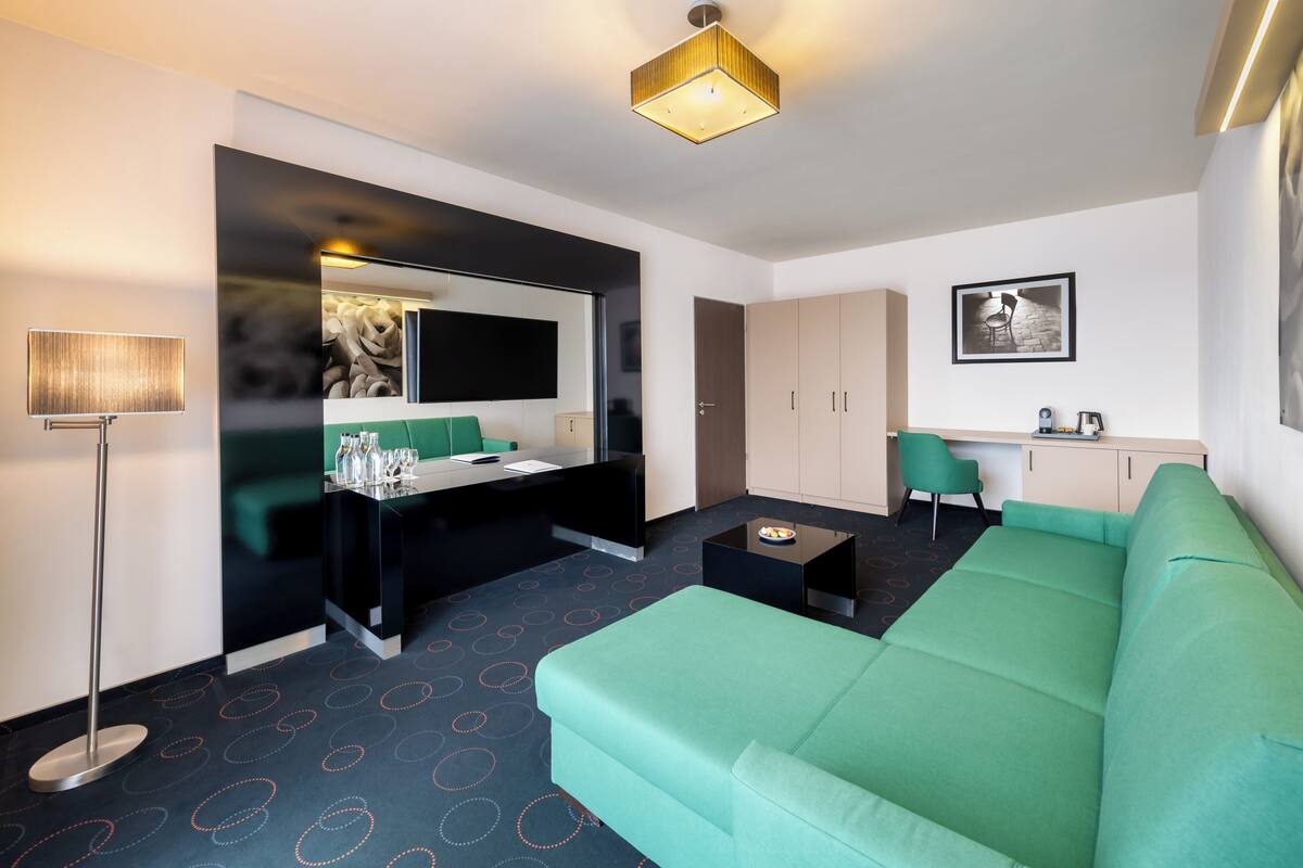 A modern hotel room with a green sofa, a small coffee table, a desk with a chair, a large mirror, and a kitchenette area.