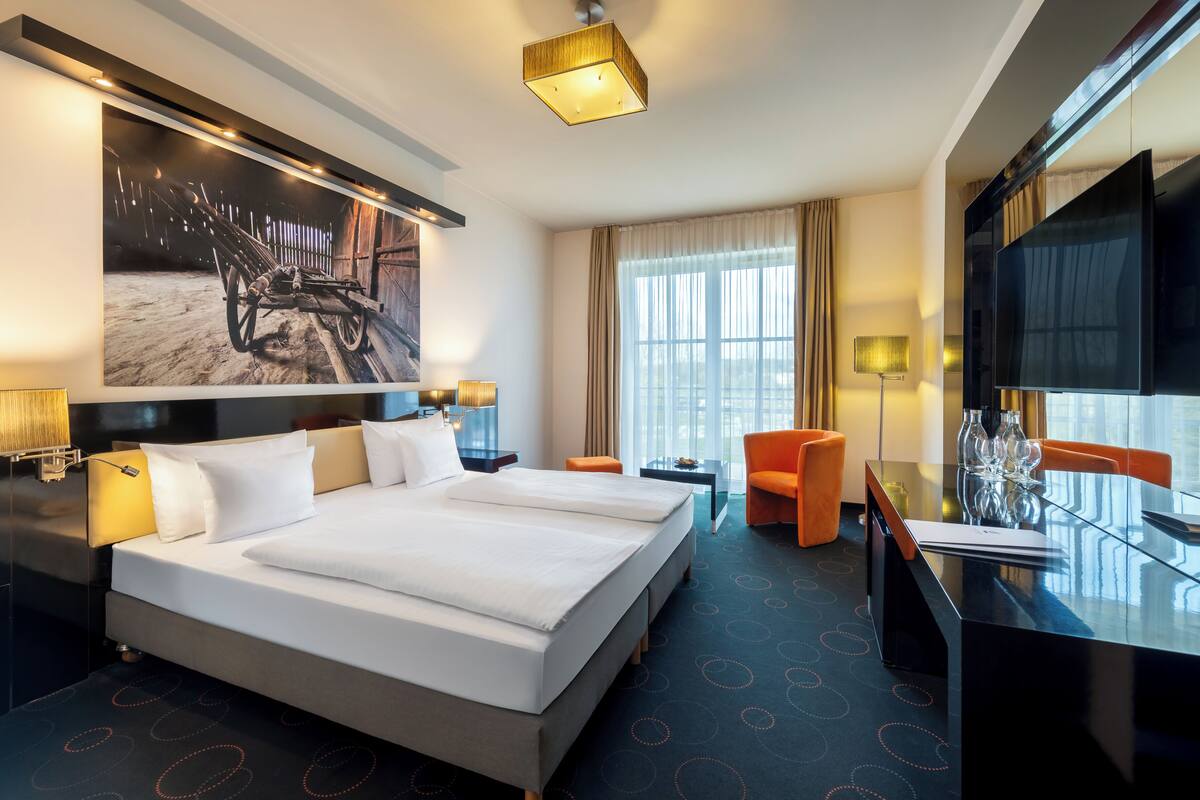 A hotel room with a large bed, a desk with a TV, a chair, and a large window.