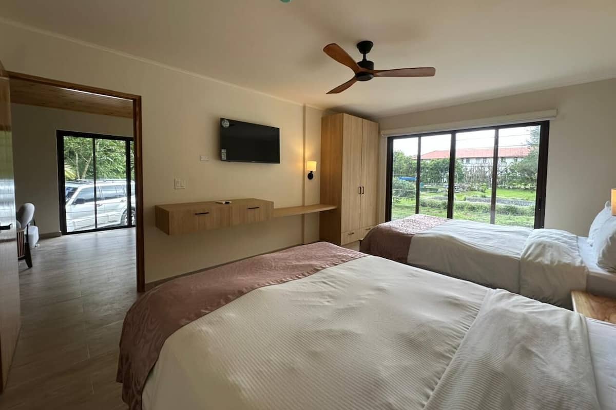 Habitación de hotel con dos camas, ventilador de techo, televisor montado en la pared y grandes ventanas con vistas al exterior.