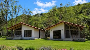 Dos edificios pequeños de una sola planta, con cimientos de piedra y techos rojos, rodeados de un césped cuidado y plantas con flores.
