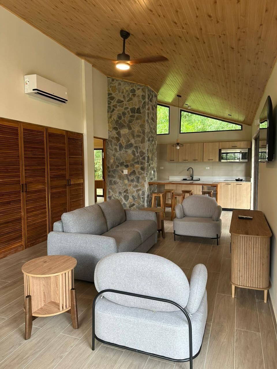 A modern living room with a stone accent wall, wooden ceiling, and a kitchen area with a stone backsplash.