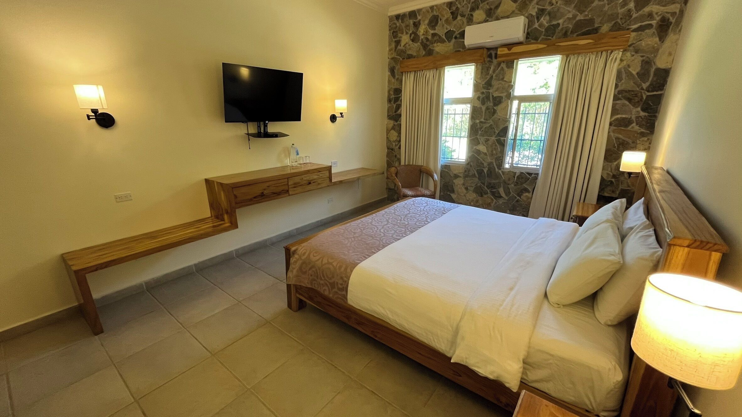 A hotel room with a bed, a television, a desk, and a stone wall.