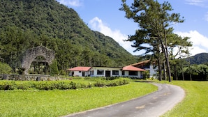 A two-story building with a red roof, surrounded by lush greenery and a mountain backdrop.