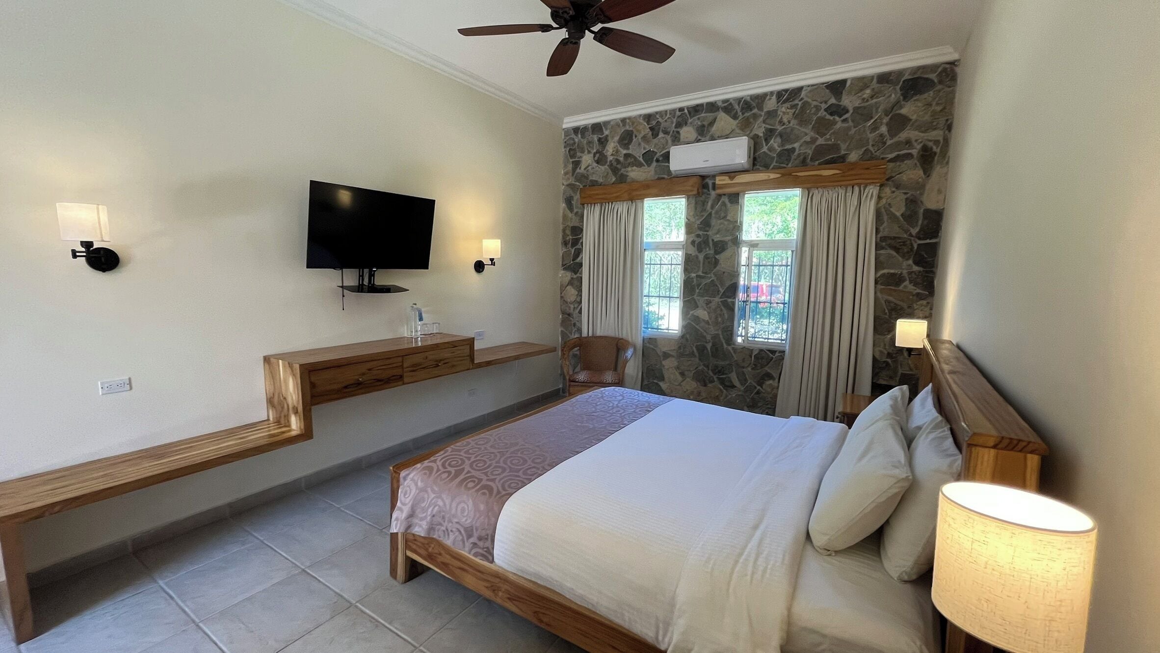 A bedroom with a bed, a television, a stone wall, and a window with curtains.