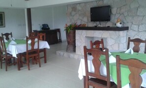 A dining area with tables set for four, wooden chairs, a stone wall, a fireplace, and a television mounted above it. - Hotel Campestre (El Valle de Anton)