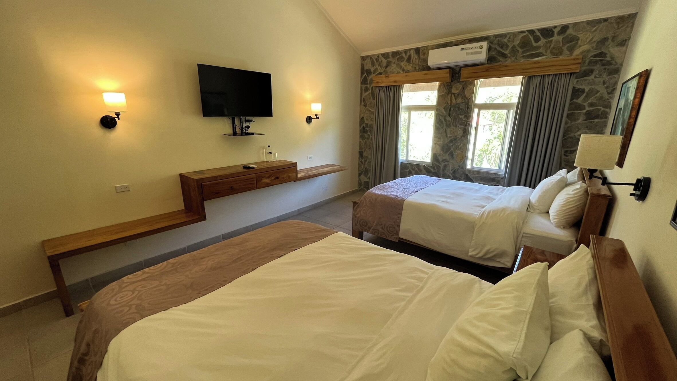 Habitación de hotel con dos camas, televisión, un muro decorativo de piedra y una ventana con cortinas.