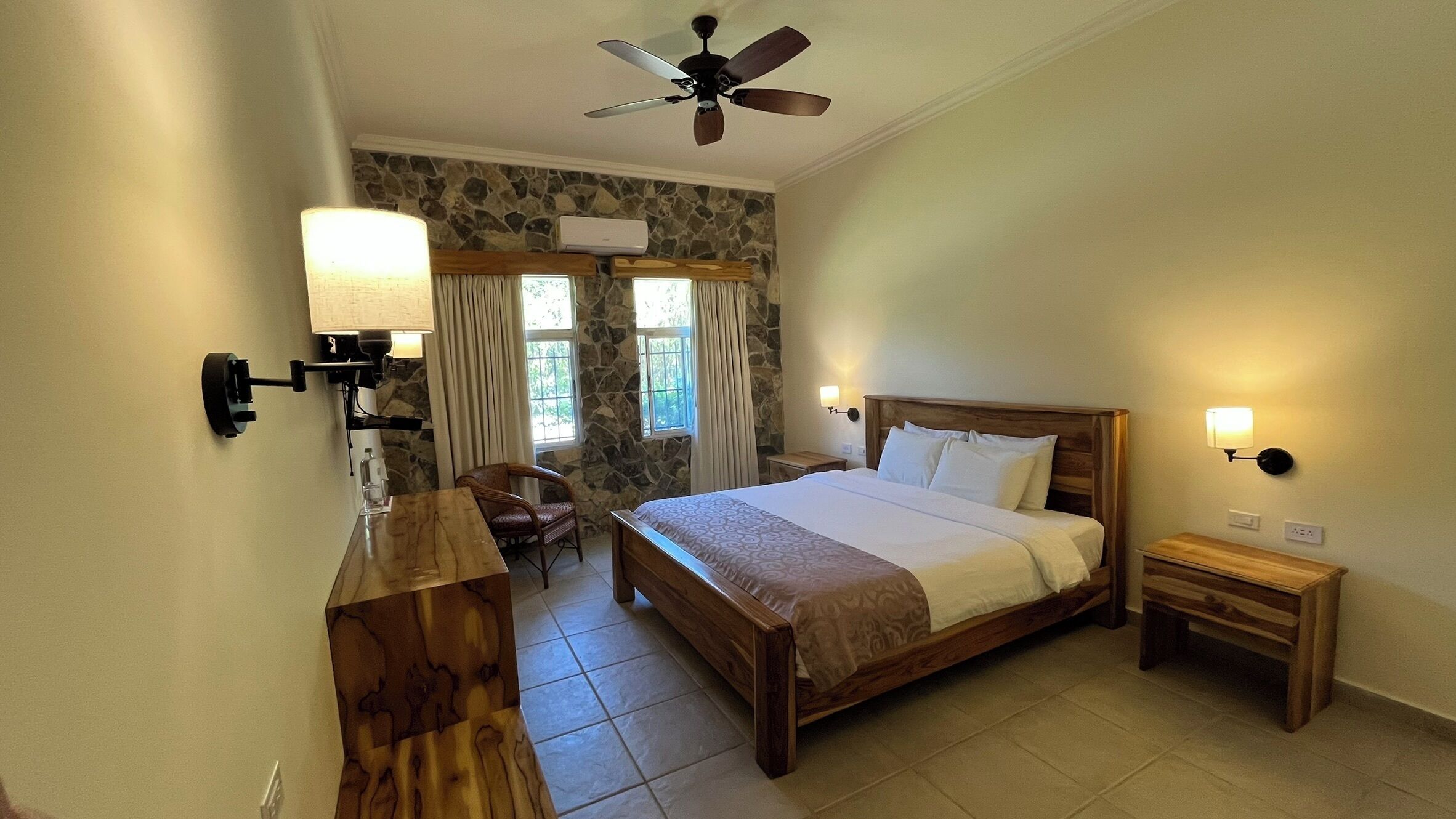 Habitación de hotel con cama, escritorio, silla, mesita de noche, lámpara de pared y ventilador de techo, además de una pared de piedra.