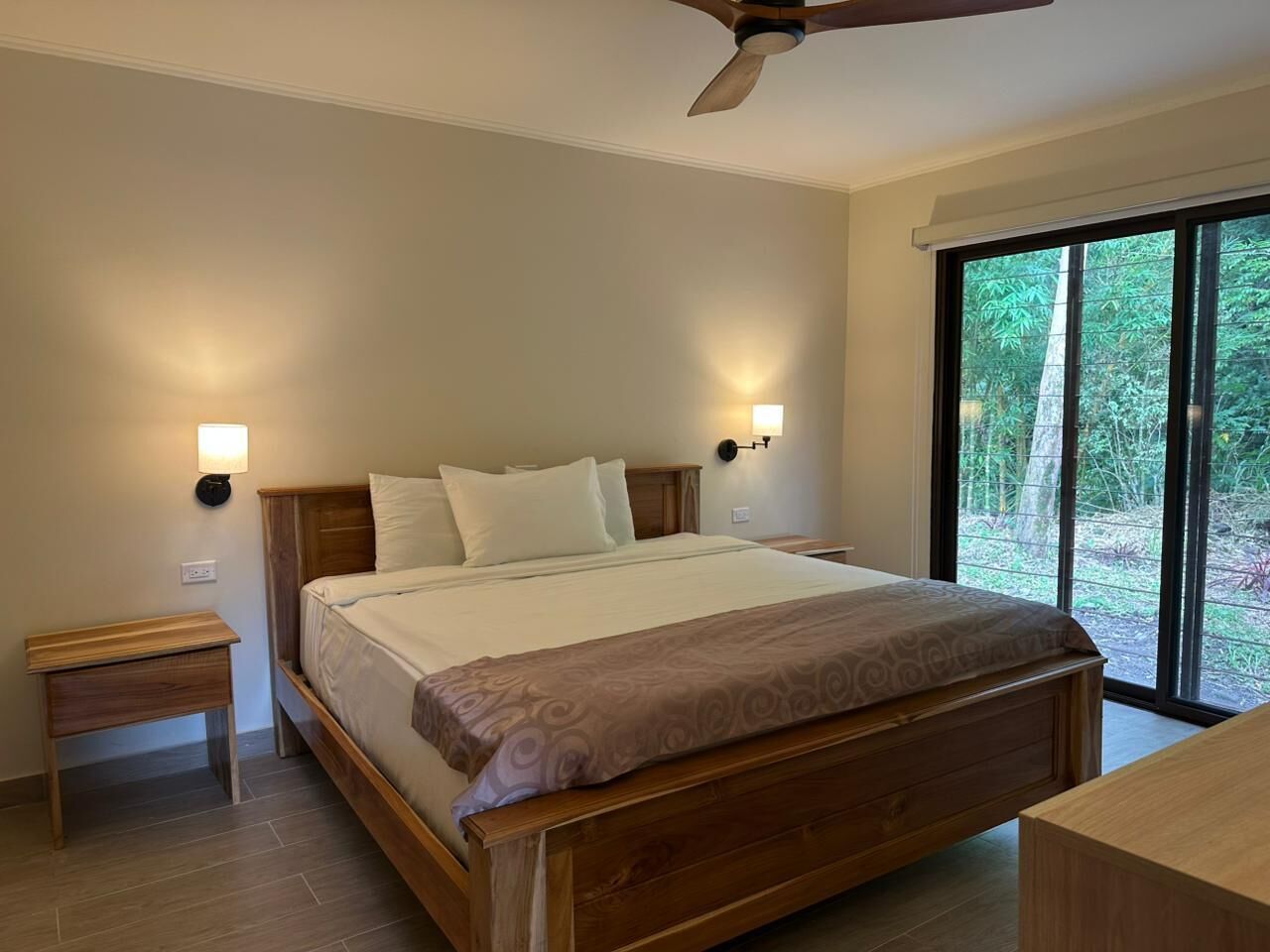 A bedroom with a bed, bedside tables, a ceiling fan, and a large window with a view of greenery.