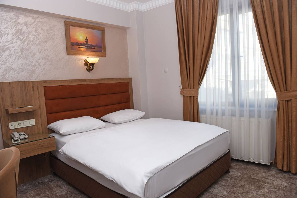Une chambre d’hôtel avec une tête de lit en bois, un lit recouvert de draps blancs, un bureau avec un téléphone et un tableau représentant un coucher de soleil accroché au mur.