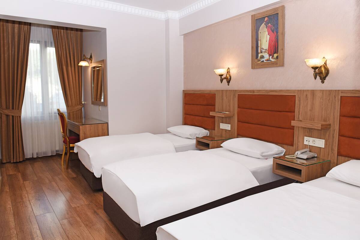 Une chambre d’hôtel avec trois lits, des têtes de lit en bois, un bureau et une chaise. Il y a des lampes fixées au mur et un tableau au-dessus des lits.