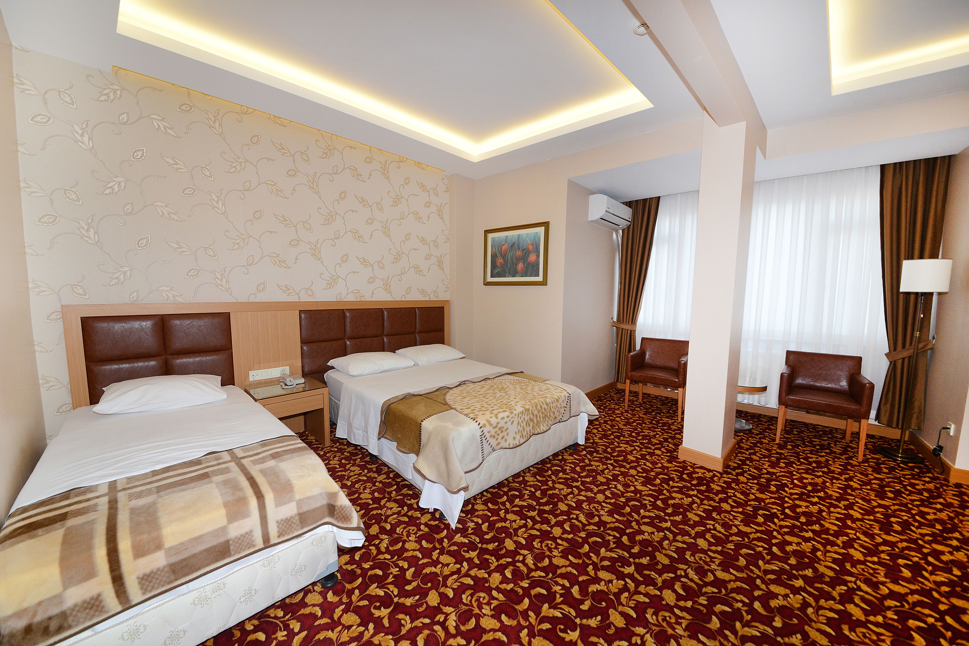 A hotel room with a large bed, a smaller bed, a desk with a chair, an air conditioning unit, and a painting on the wall.